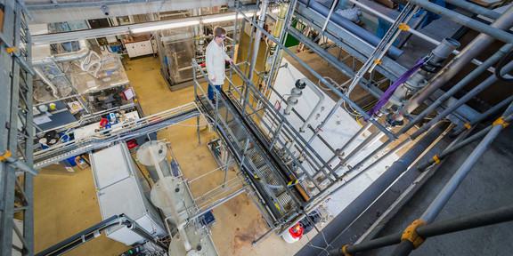 A person with a white coat stands in a laboratory with several large containers and metal pipes.
