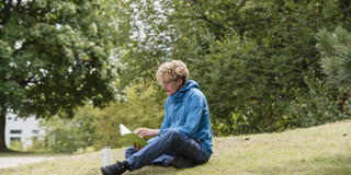 A student sits on a meadow and reads in a book.